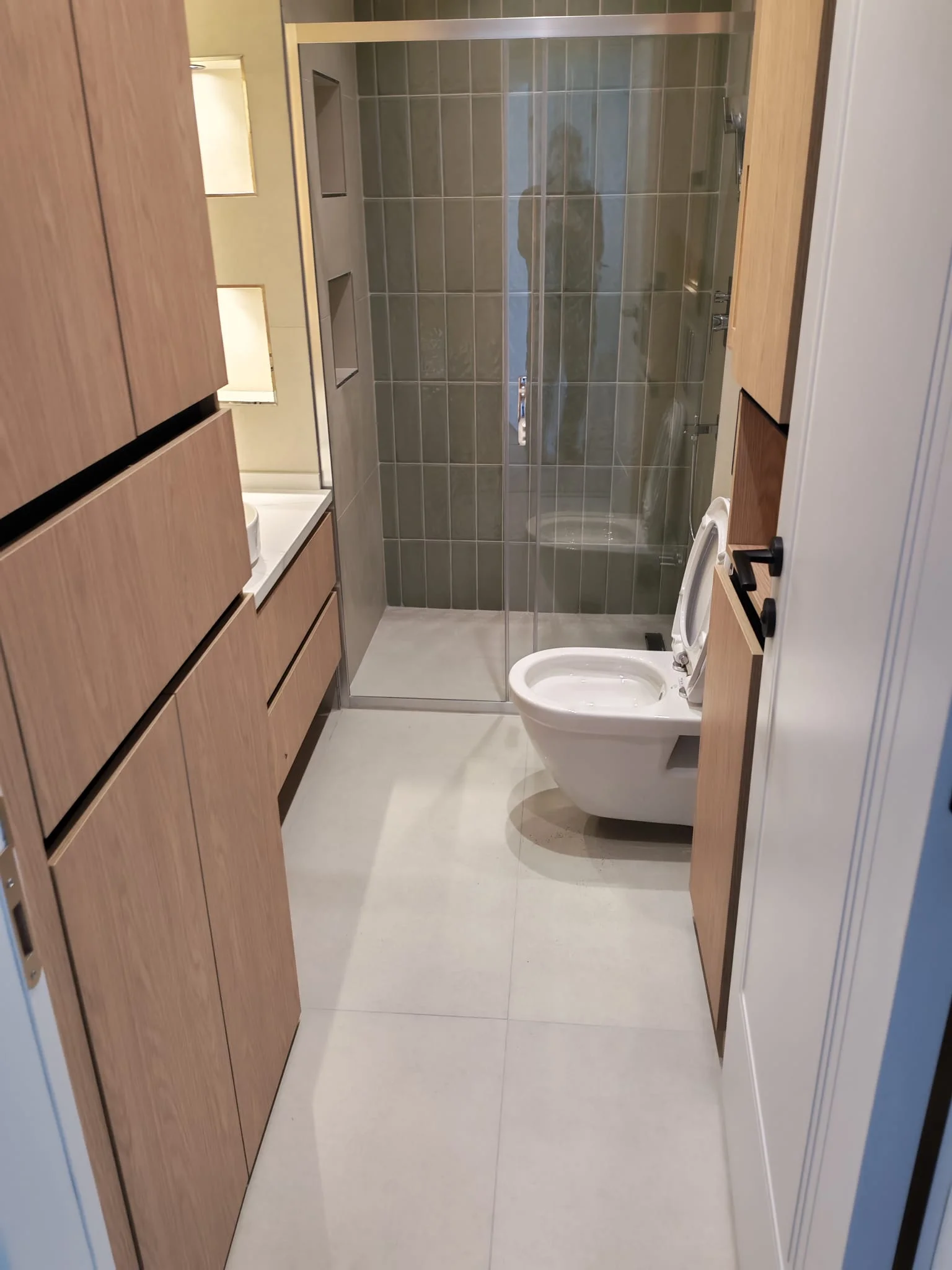 Dar banyoda ahşap lavabo dolabı, klozet, çamaşır makinesi ve cam duş bölmesi