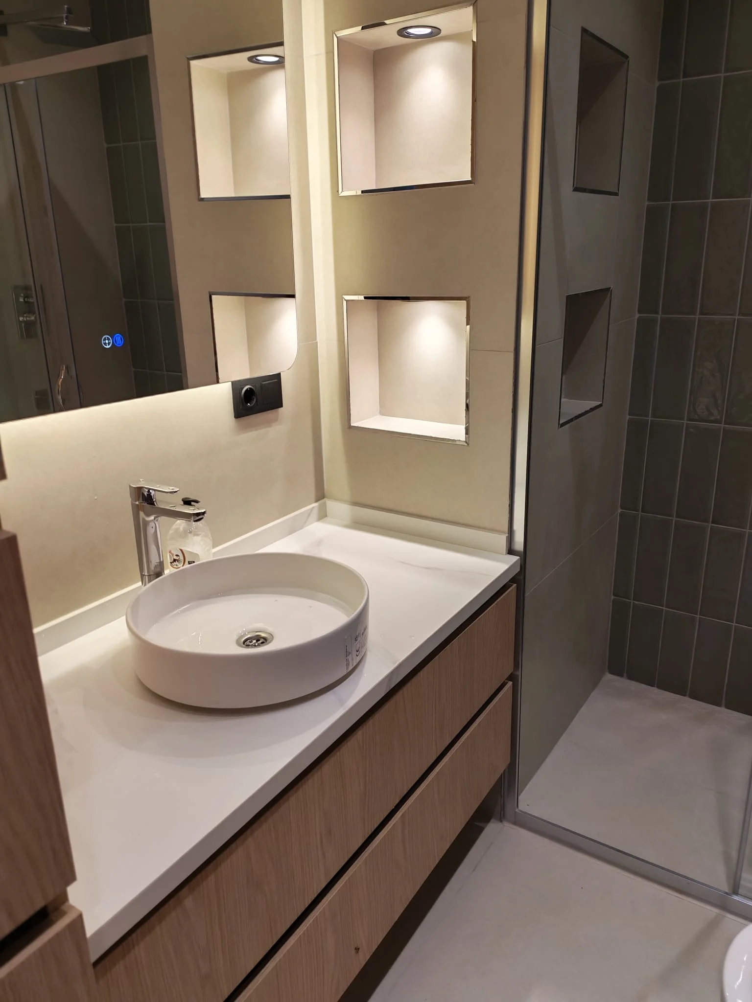 Yuvarlak çanak lavabo, aydınlatmalı ayna ve ahşap tonlu asma dolap bulunan banyo köşesi